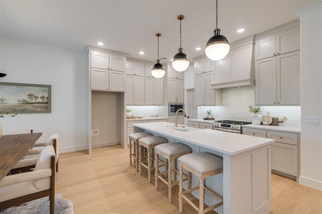 kitchen featuring premium range hood, a center island with sink, hanging light fixtures, light hardwood / wood-style floors, and stainless steel appliances