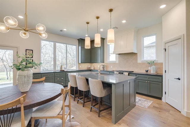 kitchen with light wood-type flooring, custom range hood, sink, pendant lighting, and an island with sink