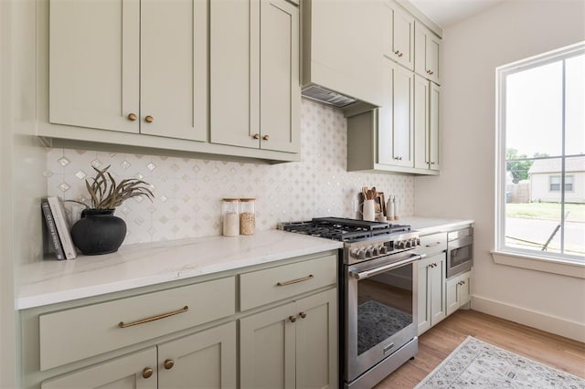 kitchen with tasteful backsplash, custom exhaust hood, stainless steel appliances, light stone countertops, and light hardwood / wood-style flooring