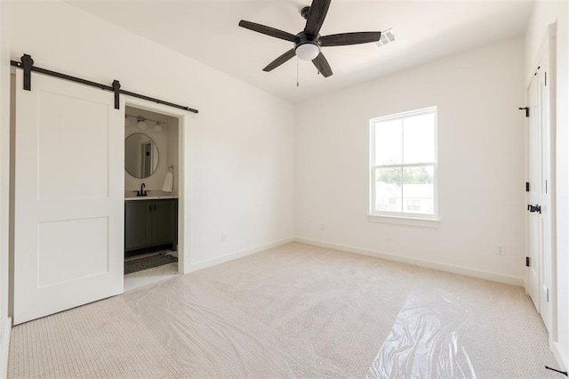 unfurnished bedroom with sink, light colored carpet, ceiling fan, a barn door, and ensuite bath