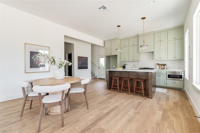 kitchen featuring tasteful backsplash, decorative light fixtures, green cabinets, an island with sink, and light hardwood / wood-style floors