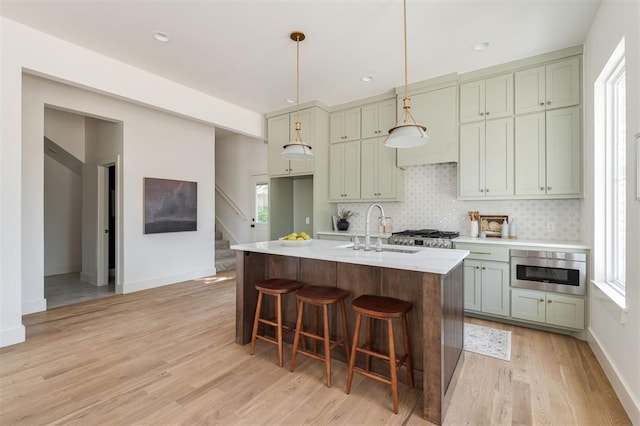 kitchen featuring sink, light hardwood / wood-style flooring, a kitchen island with sink, hanging light fixtures, and tasteful backsplash