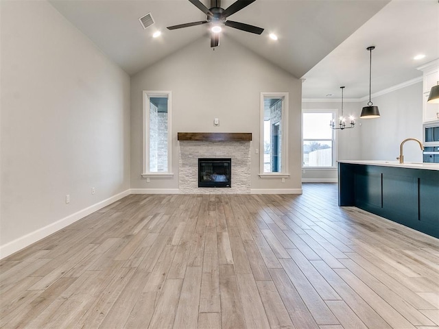 unfurnished living room with sink, a stone fireplace, light hardwood / wood-style flooring, lofted ceiling, and ceiling fan with notable chandelier