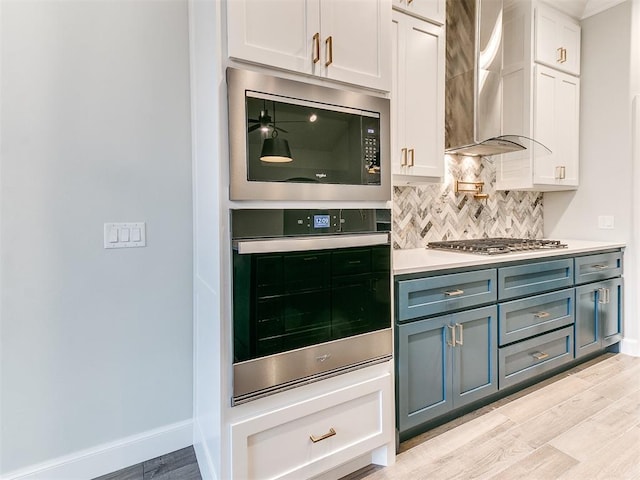 kitchen with appliances with stainless steel finishes, tasteful backsplash, wall chimney range hood, white cabinets, and light hardwood / wood-style floors