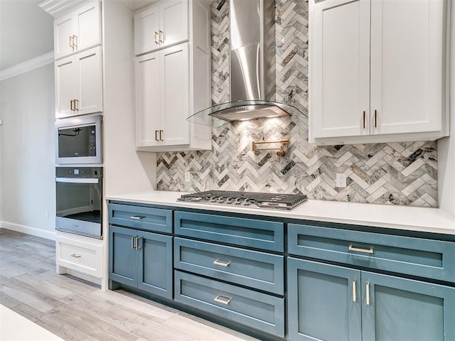 kitchen with wall chimney range hood, blue cabinets, crown molding, white cabinets, and appliances with stainless steel finishes