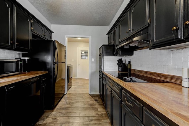 kitchen featuring butcher block counters, light wood-style flooring, under cabinet range hood, dark cabinetry, and black appliances