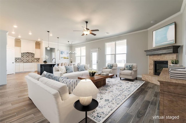 living room with crown molding, ceiling fan with notable chandelier, a fireplace, and light hardwood / wood-style flooring