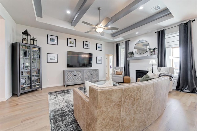 living room with a brick fireplace, ceiling fan, a raised ceiling, and light hardwood / wood-style flooring