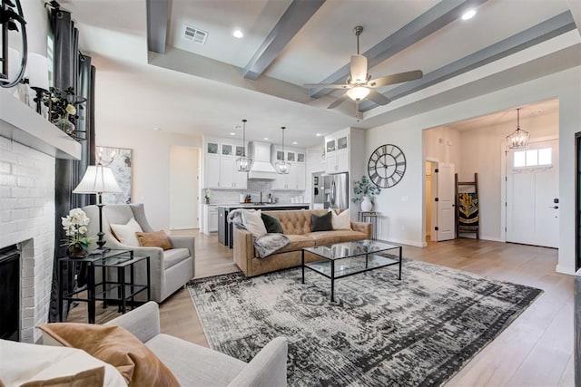 living room with ceiling fan with notable chandelier, sink, beamed ceiling, a fireplace, and light hardwood / wood-style floors