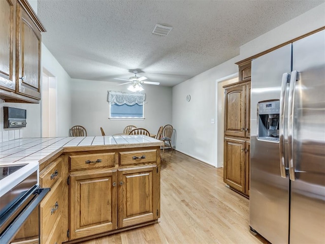 kitchen featuring stainless steel refrigerator with ice dispenser, ceiling fan, a textured ceiling, tile counters, and light hardwood / wood-style floors