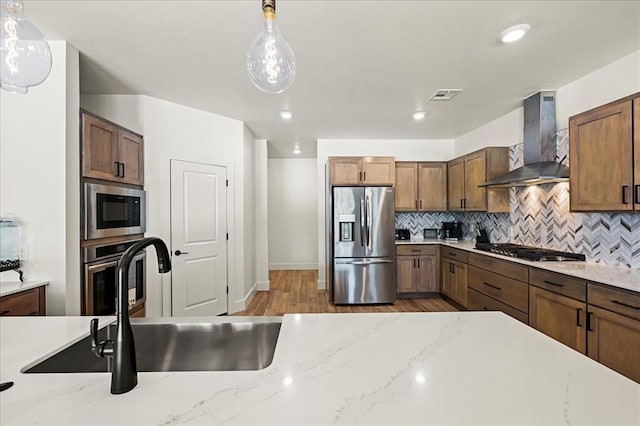 kitchen featuring wall chimney exhaust hood, light stone counters, stainless steel appliances, and hanging light fixtures