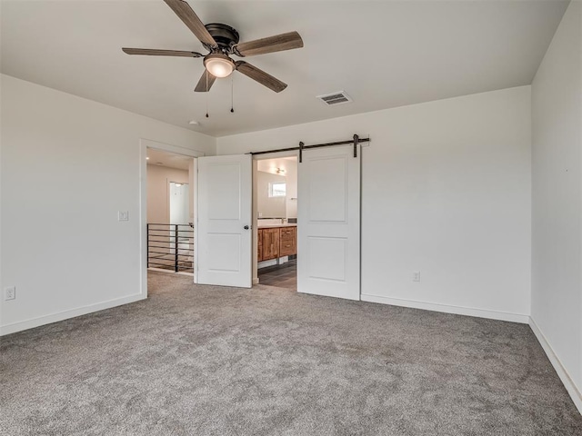 unfurnished bedroom with dark colored carpet, ceiling fan, a barn door, and ensuite bathroom