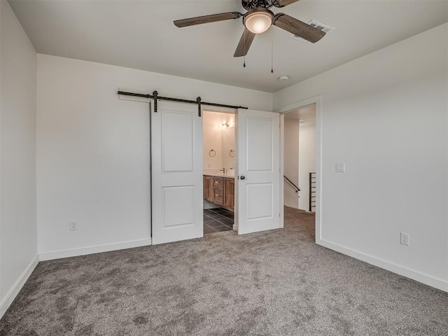 unfurnished bedroom featuring dark colored carpet, ceiling fan, a barn door, and ensuite bathroom