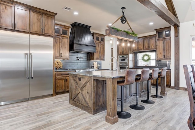 kitchen with custom exhaust hood, backsplash, a center island with sink, appliances with stainless steel finishes, and light hardwood / wood-style floors