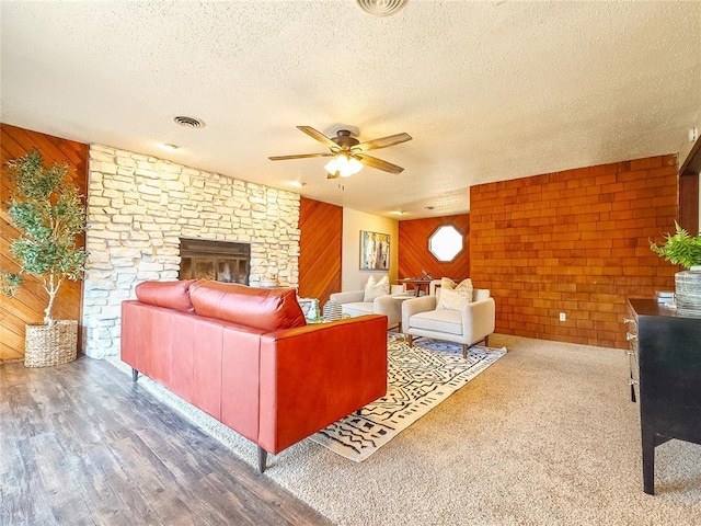 living room featuring wood walls, ceiling fan, a textured ceiling, and a stone fireplace