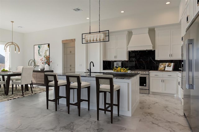 kitchen with pendant lighting, stainless steel appliances, custom range hood, white cabinets, and an island with sink