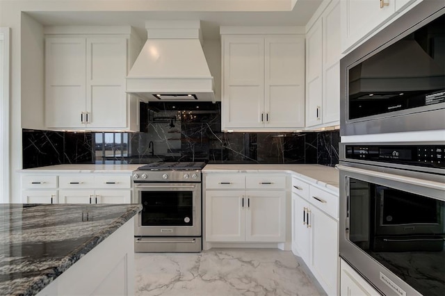 kitchen with appliances with stainless steel finishes, premium range hood, backsplash, white cabinets, and dark stone counters