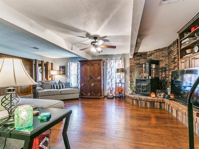 living room with a wood stove, ceiling fan, dark hardwood / wood-style flooring, beamed ceiling, and a textured ceiling