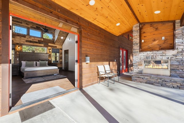 bedroom with wood walls, wooden ceiling, high vaulted ceiling, a stone fireplace, and concrete flooring