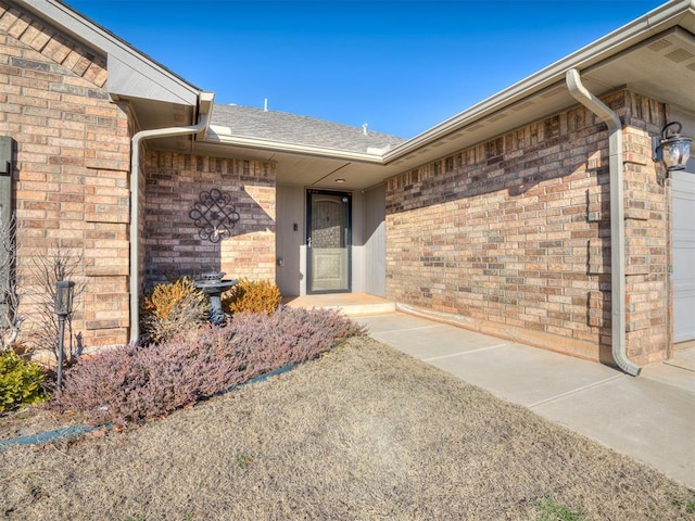 view of exterior entry with an attached garage, a shingled roof, and brick siding