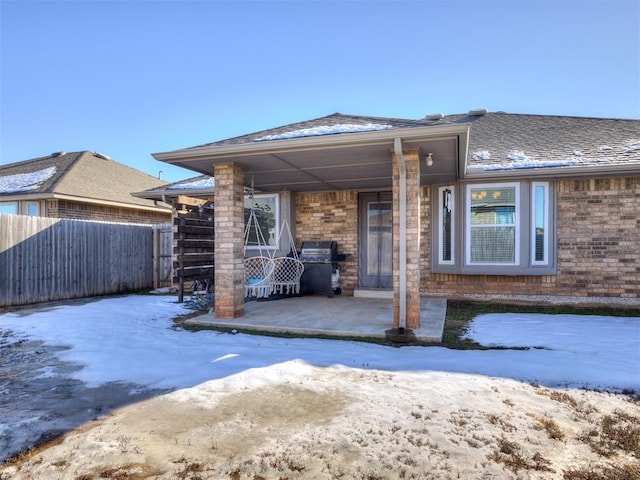 snow covered property with a patio area, a shingled roof, fence, and brick siding