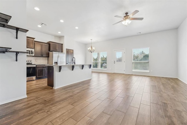 kitchen with a breakfast bar, ceiling fan with notable chandelier, light stone countertops, an island with sink, and appliances with stainless steel finishes