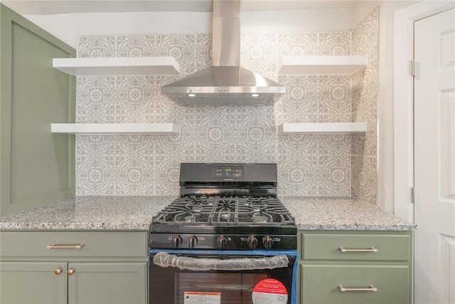 kitchen featuring black gas range, light stone countertops, wall chimney range hood, green cabinets, and decorative backsplash