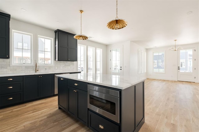 kitchen featuring light wood finished floors, stainless steel microwave, hanging light fixtures, decorative backsplash, and a sink