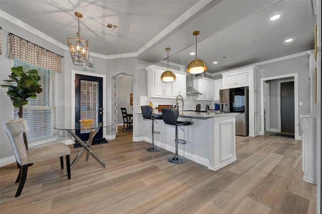 kitchen featuring stainless steel refrigerator with ice dispenser, wall chimney exhaust hood, decorative backsplash, decorative light fixtures, and white cabinetry