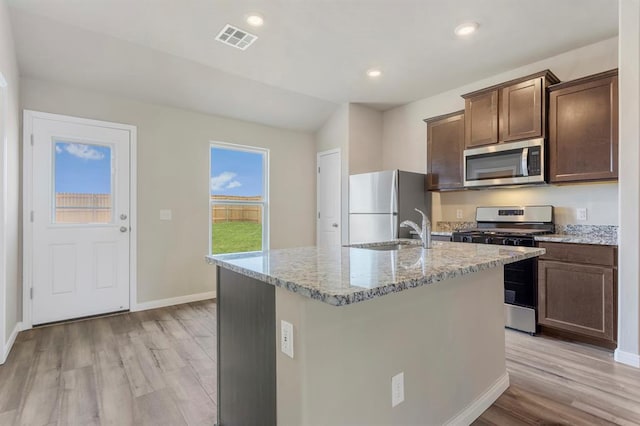 kitchen with a center island with sink, sink, light wood-type flooring, light stone counters, and stainless steel appliances