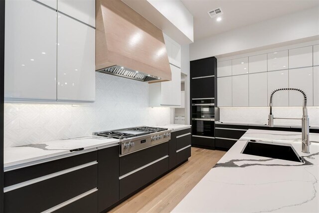 kitchen with light stone countertops, stainless steel gas cooktop, sink, wall chimney range hood, and white cabinets