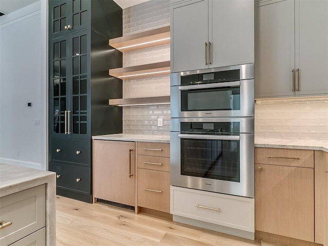 kitchen featuring light stone countertops, light brown cabinetry, tasteful backsplash, double oven, and light hardwood / wood-style floors