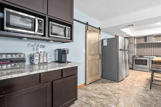 kitchen with a barn door, a textured ceiling, appliances with stainless steel finishes, light stone counters, and dark brown cabinetry