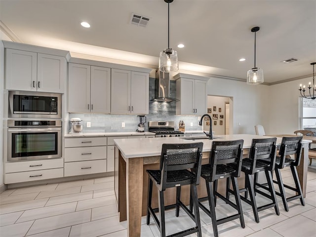 kitchen with pendant lighting, wall chimney exhaust hood, sink, an island with sink, and stainless steel appliances