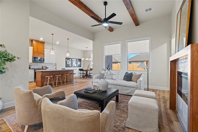 living area featuring beamed ceiling, ceiling fan with notable chandelier, visible vents, and light wood finished floors