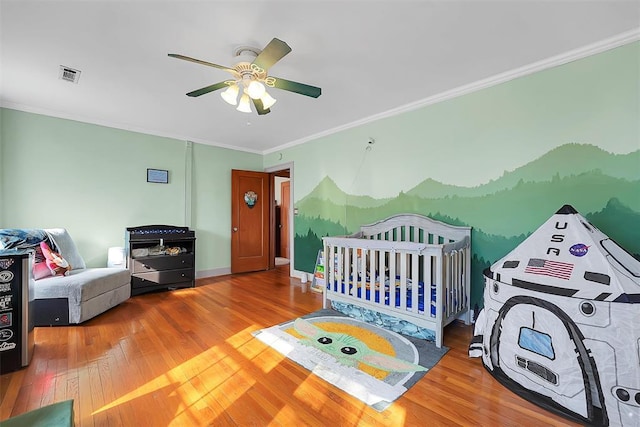 bedroom featuring a crib, crown molding, wood-type flooring, and ceiling fan