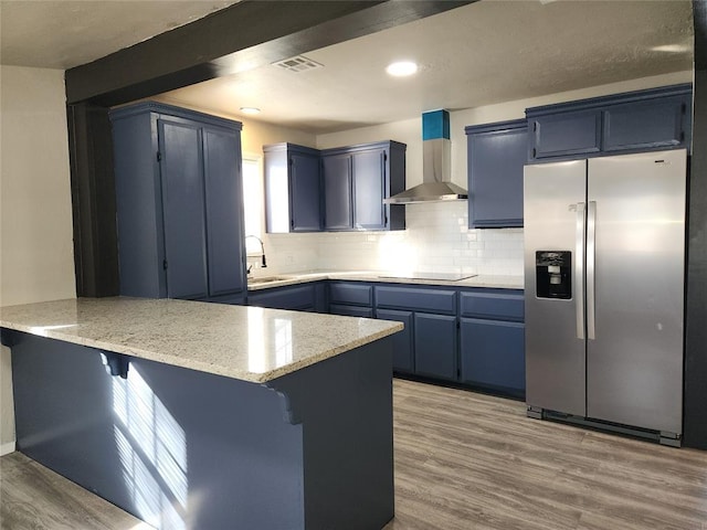 kitchen with kitchen peninsula, stainless steel fridge, blue cabinets, and wall chimney exhaust hood