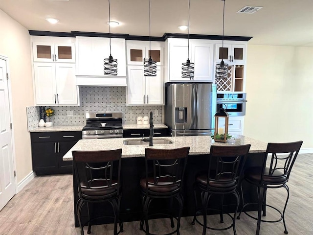 kitchen featuring a kitchen island with sink, visible vents, hanging light fixtures, appliances with stainless steel finishes, and glass insert cabinets