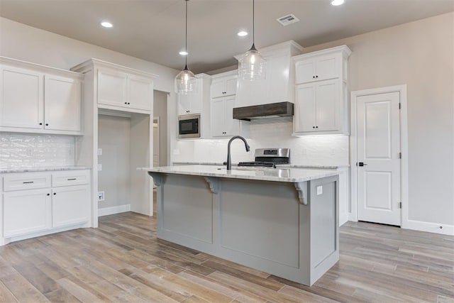 kitchen with built in microwave, an island with sink, decorative light fixtures, stainless steel range oven, and white cabinetry