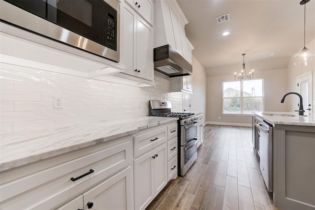 kitchen with stainless steel appliances, wall chimney range hood, sink, a notable chandelier, and white cabinets