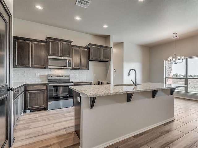 kitchen featuring a kitchen island with sink, an inviting chandelier, sink, light stone countertops, and appliances with stainless steel finishes