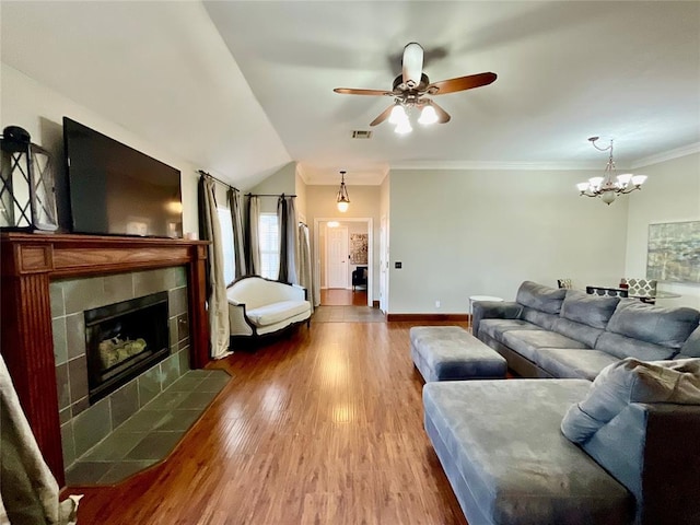 living room featuring wood-type flooring, lofted ceiling, a tiled fireplace, ceiling fan with notable chandelier, and ornamental molding