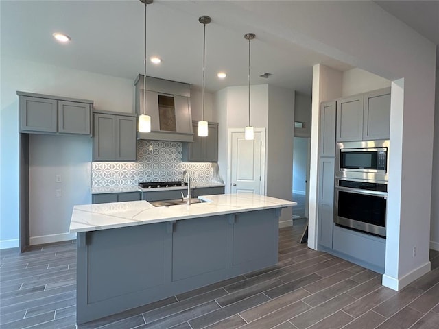 kitchen with premium range hood, light stone countertops, an island with sink, tasteful backsplash, and stainless steel appliances