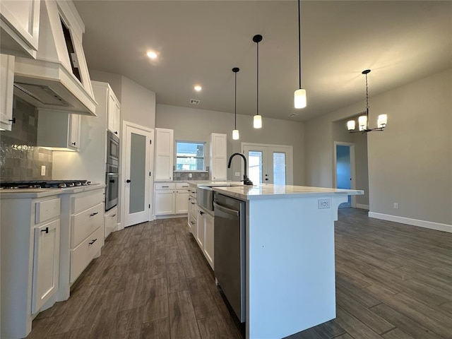 kitchen featuring premium range hood, appliances with stainless steel finishes, an island with sink, white cabinets, and decorative light fixtures