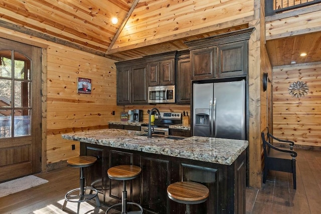 kitchen with wood walls, light stone countertops, an island with sink, stainless steel appliances, and dark brown cabinets