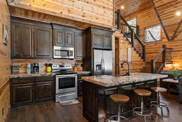 kitchen with wooden walls, appliances with stainless steel finishes, a kitchen island with sink, light stone counters, and sink