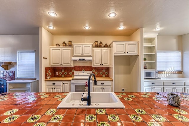 kitchen with electric range, tile countertops, under cabinet range hood, white cabinetry, and a sink