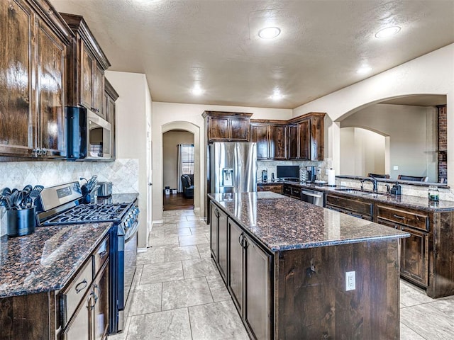 kitchen featuring dark brown cabinets, a center island, appliances with stainless steel finishes, arched walkways, and a sink