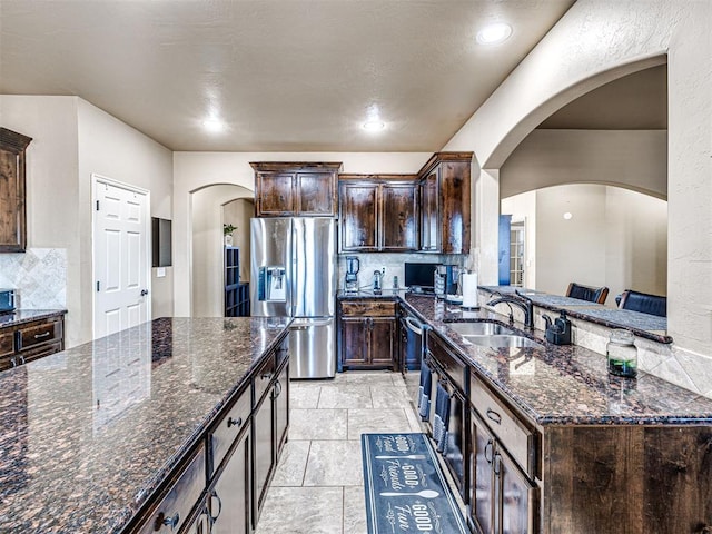 kitchen with dark brown cabinetry, dark stone counters, decorative backsplash, appliances with stainless steel finishes, and a sink