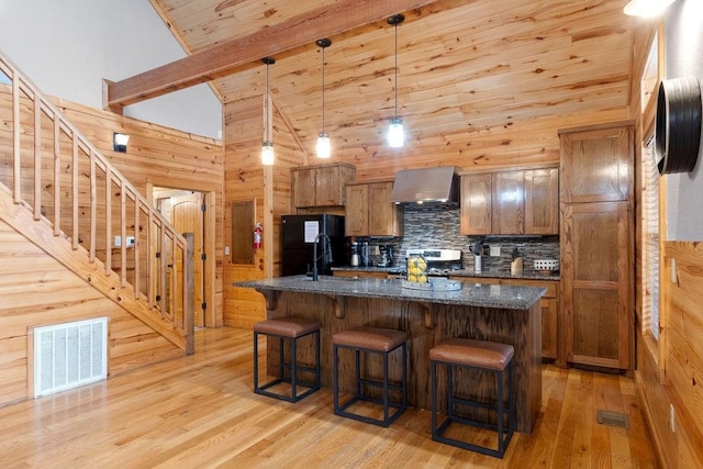 kitchen featuring wall chimney range hood, dark stone countertops, hanging light fixtures, a kitchen island with sink, and high vaulted ceiling